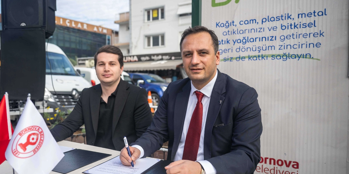 Bornova Belediyesi'nin Atık Bank Projesi açılışında, lacivert takım elbiseli ve kırmızı kravatlı bir yetkili masada oturarak imza atıyor, yanında ise siyah ceketli bir başka kişi ona eşlik ediyor. Masanın üzerinde Bornova Belediyesi logolu küçük bir Türk bayrağı bulunuyor. Arkalarındaki panelde 