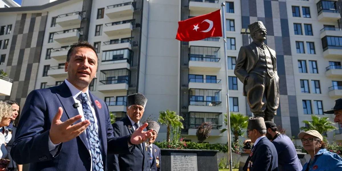 Modern bir apartman binasının önünde, elleriyle konuşan bir konuşmacı ve yanında geleneksel kalpaklı gaziler yer alıyor. Arka planda, üzerinde Türk bayrağının dalgalandığı bir Atatürk heykeli bulunuyor. Konuşmacı lacivert takım elbise ve desenli bir kravat giymiş, etrafında ise madalyalı gaziler ve katılımcılar dikkat çekiyor.
