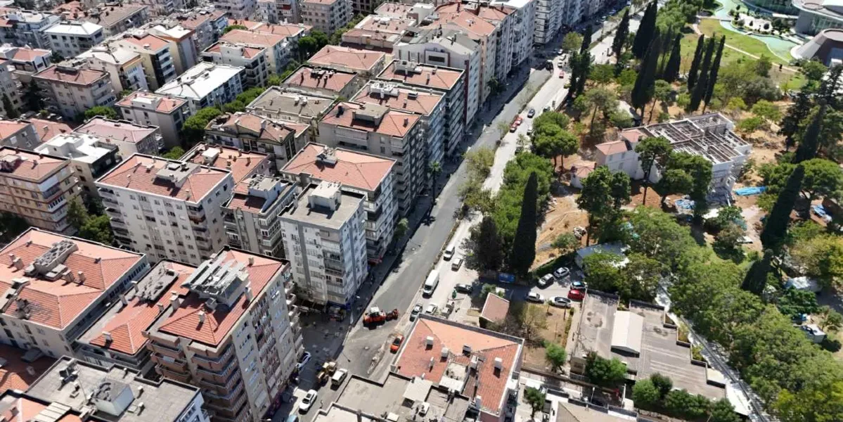 Bornova'da çok katlı apartmanların bulunduğu yoğun yerleşim bölgesinde, asfaltlama çalışması yapılan bir caddeyi gösteren kuş bakışı görünüm. Cadde üzerinde iş makineleri ve çalışanlar yer alırken, yolun bir tarafında ağaçlık bir alan ve park bölgesi uzanıyor.