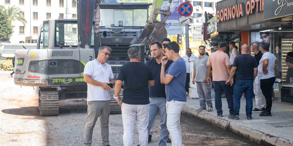 Mustafa Kemal Caddesi'ndeki yol yenileme çalışmaları sırasında, arkalarında büyük bir iş makinesi bulunan bir grup erkek, kaldırım kenarında durmuş sohbet ediyor. Sağ tarafta Bornova Optik tabelası ve dükkan vitrini görülüyor.