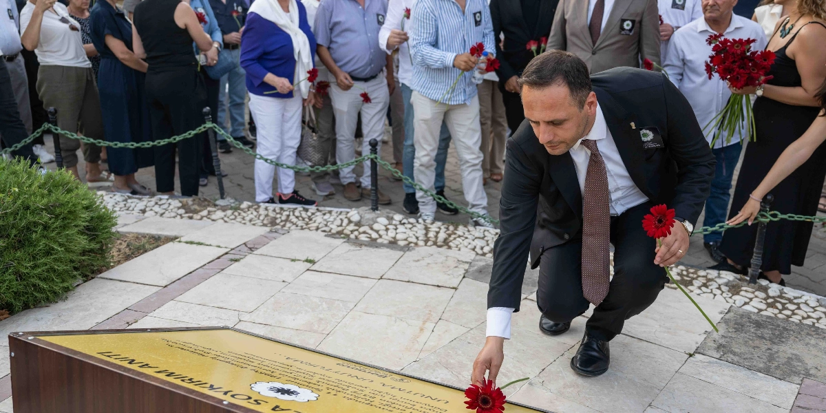 Takım elbiseli bir erkek, Bornova'daki Srebrenitsa Soykırımı Anıtı'na kırmızı bir çiçek bırakıyor. Anma törenine katılan diğer kişiler de ellerinde kırmızı çiçeklerle anıtın çevresinde duruyor. Anıtın üzerinde 