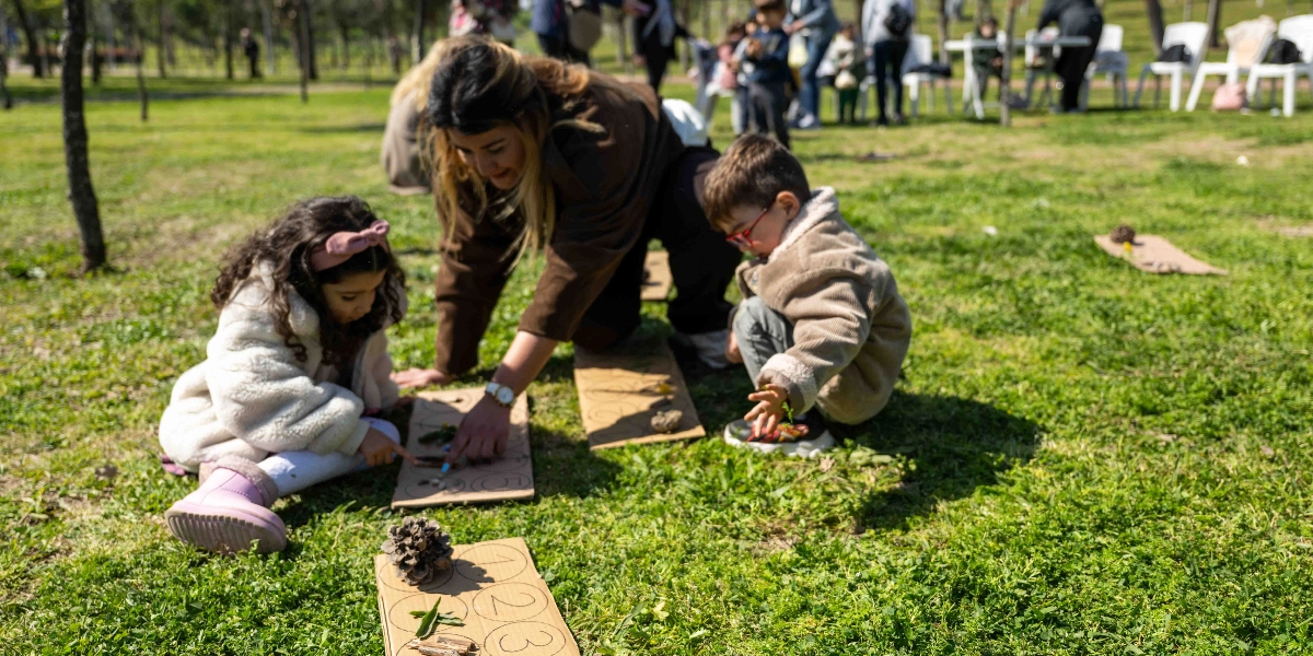 Açık havada, yeşil bir alanda diz çökmüş bir öğretmen, biri kız diğeri erkek iki çocukla birlikte yere serilmiş kartonlar üzerinde doğa materyalleriyle oyun oynuyor. Çocuklar ve öğretmen, üzerinde rakamlar ve daireler çizili kartonların içine kozalak, dal parçası ve yaprak gibi nesneleri yerleştirerek sayı sayma pratiği yapıyor. Arka planda ağaçlar ve etkinlikteki diğer katılımcılar bulanık bir şekilde görülüyor.