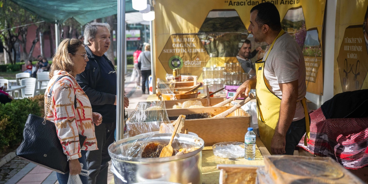 Açık havada kurulan bir festival standında, sarı önlüklü bir satıcı, tezgahındaki bal ürünleri hakkında orta yaşlı bir çifte bilgi veriyor. Tezgahın üzerinde ahşap kasalar içinde petek ballar ve çeşitli bal ürünleri sergileniyor. Arka planda 