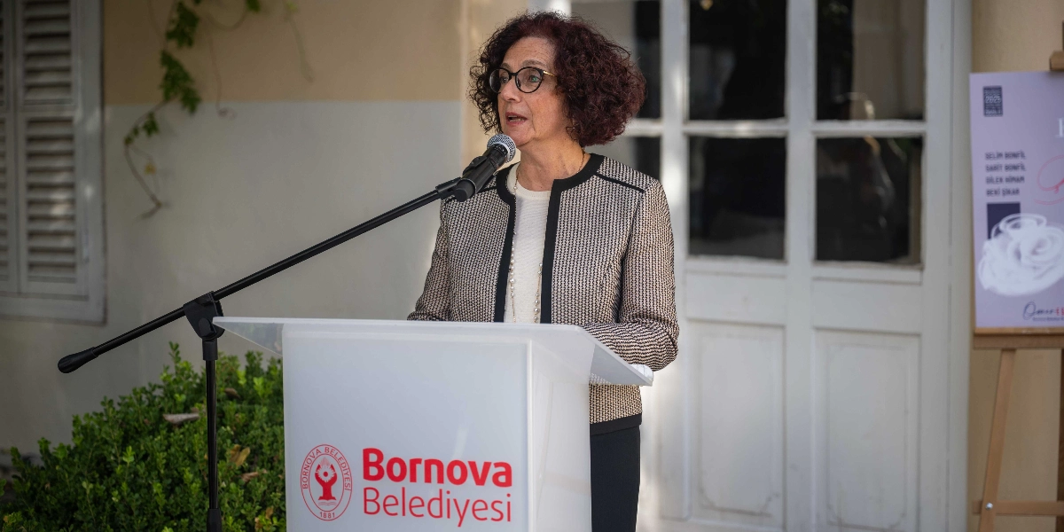 Kıvırcık kızıl saçlı, gözlüklü bir kadın, üzerinde Bornova Belediyesi logosu bulunan bir kürsüde mikrofon başında konuşma yapıyor. Kadın, krem rengi bir bluz üzerine desenli bir ceket giymiş. Arka planda beyaz bir kapı ve sağ tarafta üzerinde sergi bilgilerinin yer aldığı bir afişin bir kısmı görünüyor.