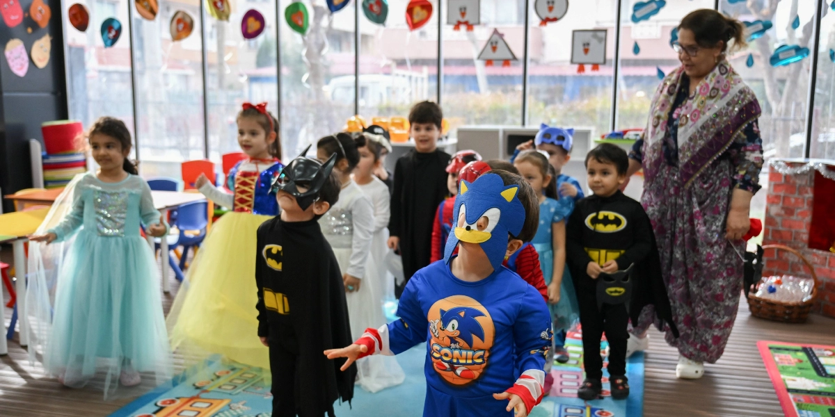 Çeşitli süper kahraman ve masal karakteri kostümleri giymiş çocukların bulunduğu bir anaokulu sınıfında, ön planda Sonic kostümlü bir çocuk ve Batman maskeli bir çocuk yer alıyor. Arkalarında Pamuk Prenses kostümlü bir kız çocuğu, diğer çocuklar ve onlara eşlik eden yetişkin bir kadın bulunuyor. Sınıfın camlarında süslemeler asılı ve zemin çocuk oyun halısıyla kaplı.