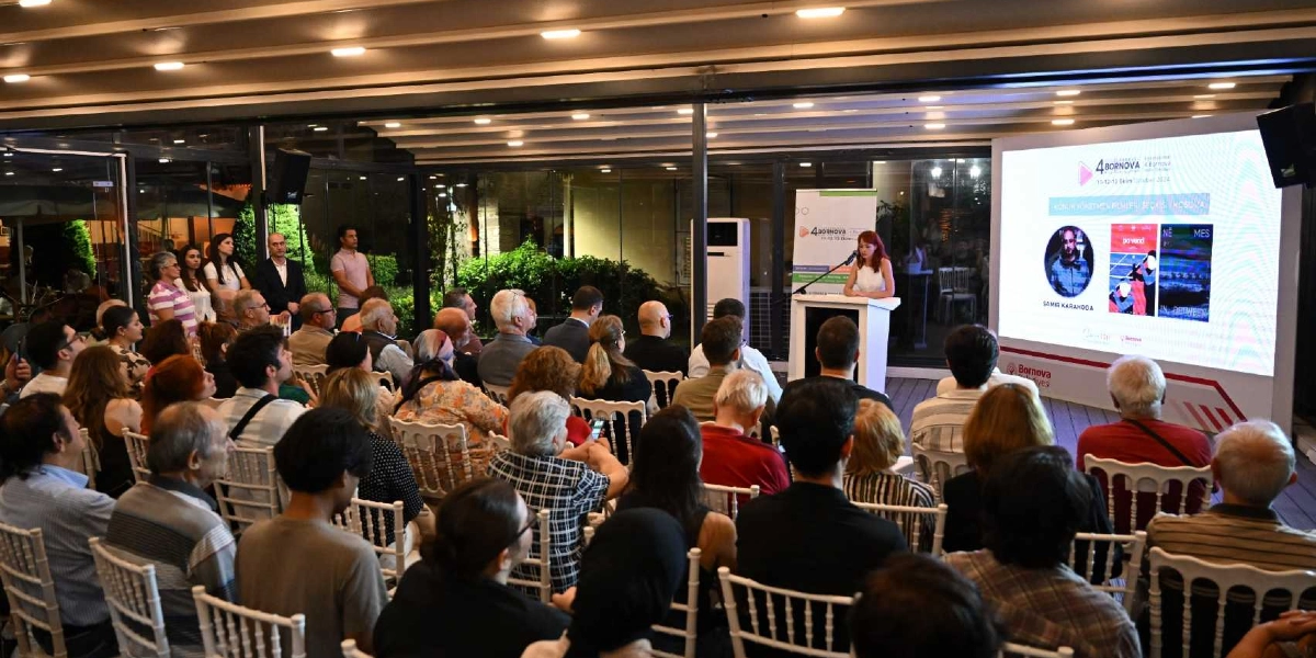 Kapalı bir mekanda düzenlenen Bornova Kısa Film Günleri etkinliğinde, kürsüde konuşma yapan bir kadın ve onu dinleyen kalabalık bir izleyici grubu yer alıyor. Sahnedeki büyük ekranda etkinliğin logosu, tarihleri ve konuk yönetmen Samir Karahoda'nın fotoğrafı ile film afişleri görünüyor. İzleyiciler beyaz sandalyelerde oturarak sunumu takip ediyor.