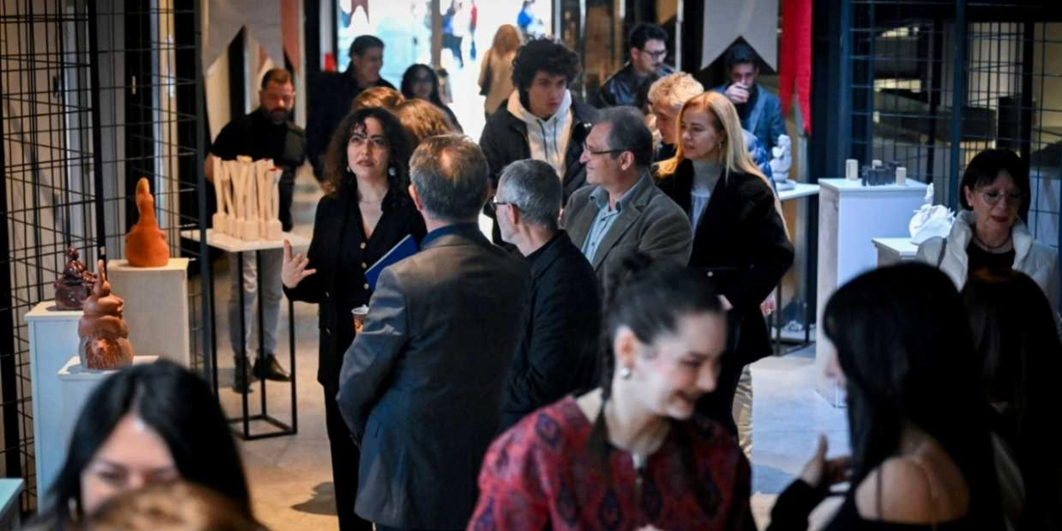 Kapalı bir mekanda düzenlenen sanat sergisinde, ziyaretçiler stantlar üzerindeki heykelleri inceliyor ve birbirleriyle sohbet ediyor. Ön planda, koyu renkli kıyafetler giymiş bir kadın, elindeki mavi dosyayla bir gruba hitap ederken, çevresinde farklı yaş gruplarından insanlar sergilenen eserleri gözlemliyor. Mekanın genelinde modern bir atmosfer hakim ve sergi alanında çeşitli figüratif heykeller yer alıyor.