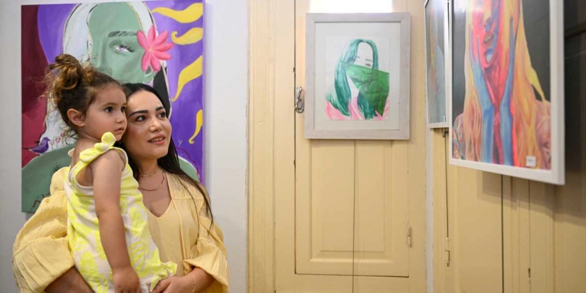 Sarı kıyafetli bir kadın, kucağındaki küçük kız çocuğuyla birlikte bir sanat galerisinde duvarda asılı olan renkli tabloları inceliyor. Kadın ve çocuk, sol tarafta yer alan büyük, mor ve yeşil tonlarındaki portre çalışmasına ve sağ tarafta çerçevelenmiş diğer eserlere bakıyor.