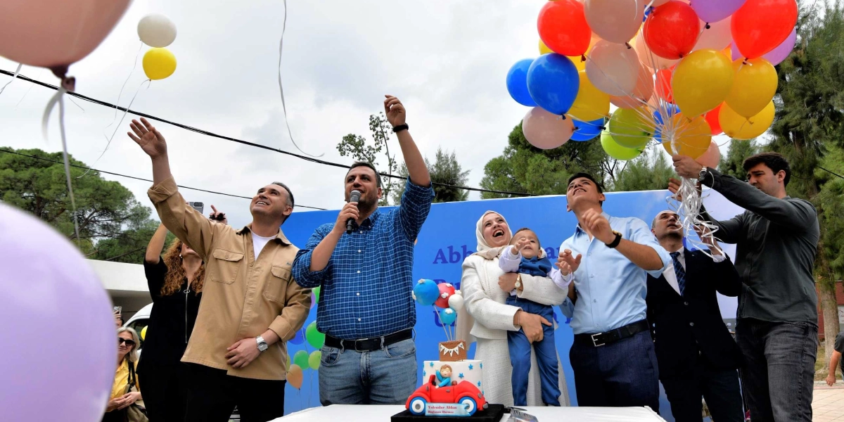 Açık havada, mavi bir fon önünde duran bir grup insan, ellerindeki renkli balonları gökyüzüne bırakıyor. Mavi ekose gömlekli bir adam elinde mikrofonla konuşurken, yanında beyaz ceketli bir kadın kucağında küçük bir çocukla duruyor. Önlerindeki masada üzerinde 