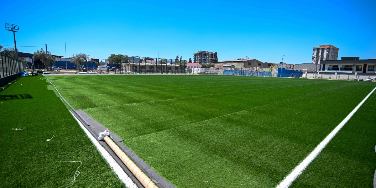 Açık havada, güneşli bir günde çekilmiş, yeni yapılmış suni çim kaplı futbol sahası. Saha çizgileri belirgin bir şekilde görülürken, kenarda henüz döşenmemiş çim rulosu ve inşaat malzemeleri yer alıyor. Arka planda ise Bornova'daki yerleşim yerleri ve binalar bulunuyor.