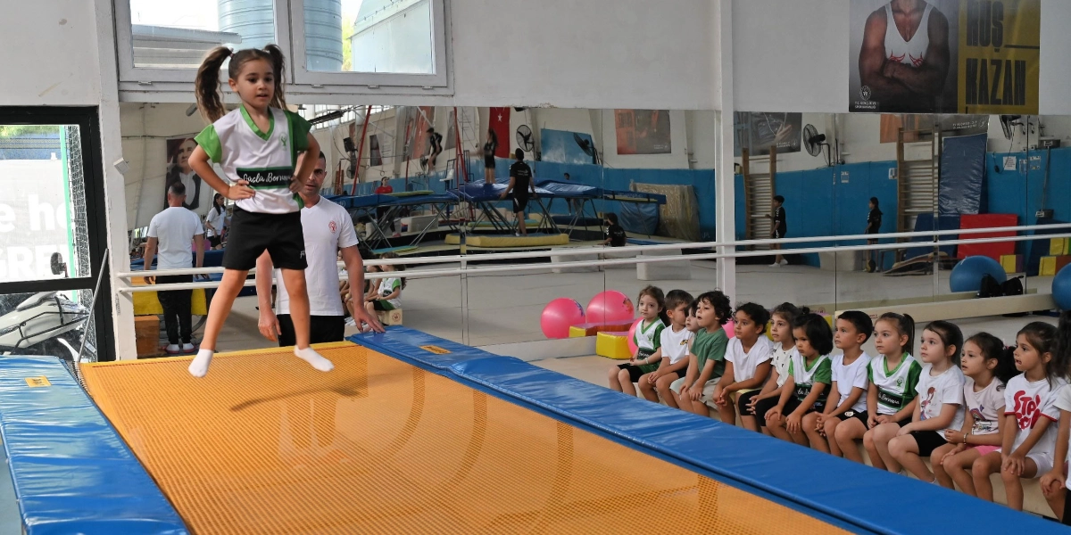 Kapalı bir spor salonunda, antrenörünün gözetiminde trambolinde zıplayan küçük bir kız çocuğu ve onu izleyen bir grup çocuk. Kız çocuğu, üzerinde 