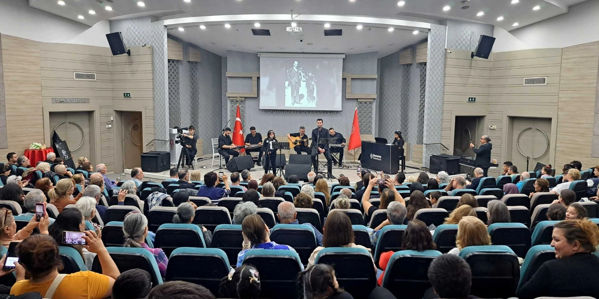 Bornova Belediyesi'ne ait bir konferans salonunda düzenlenen etkinlikte, sahnede müzik grubu ve solist performans sergiliyor. Sahnenin arka planındaki dev ekranda Mustafa Kemal Atatürk'ün siyah beyaz bir fotoğrafı yansıtılıyor. Sahnenin iki yanında Türk bayrağı ve Bornova Belediyesi'nin bayrağı yer alıyor. Salonu dolduran izleyiciler, sahnedeki performansı ilgiyle izliyor ve bazıları cep telefonlarıyla kayıt alıyor. Salonun genelinde modern bir mimari ve oturma düzeni hakim.