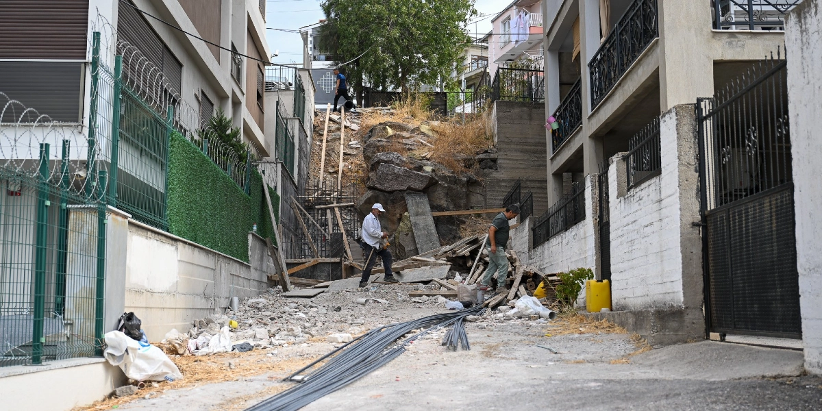 Bornova'da iki apartman arasındaki eğimli arazide, kayalık bir zemin üzerinde devam eden merdiven inşaatı çalışması. İnşaat alanında çalışan iki işçi, etrafa yayılmış tahta kalıplar, demir çubuklar ve molozlar arasında hareket ediyor. Arka planda yokuşun üst kısmında bir başka kişi daha görülüyor.