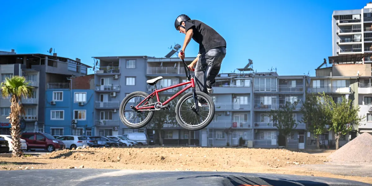 Kask takan bir genç, kırmızı bir BMX bisikletle havada zıplayarak akrobatik bir hareket sergiliyor. Arka planda şehir yerleşimi, apartmanlar ve park halindeki araçlar bulunuyor.