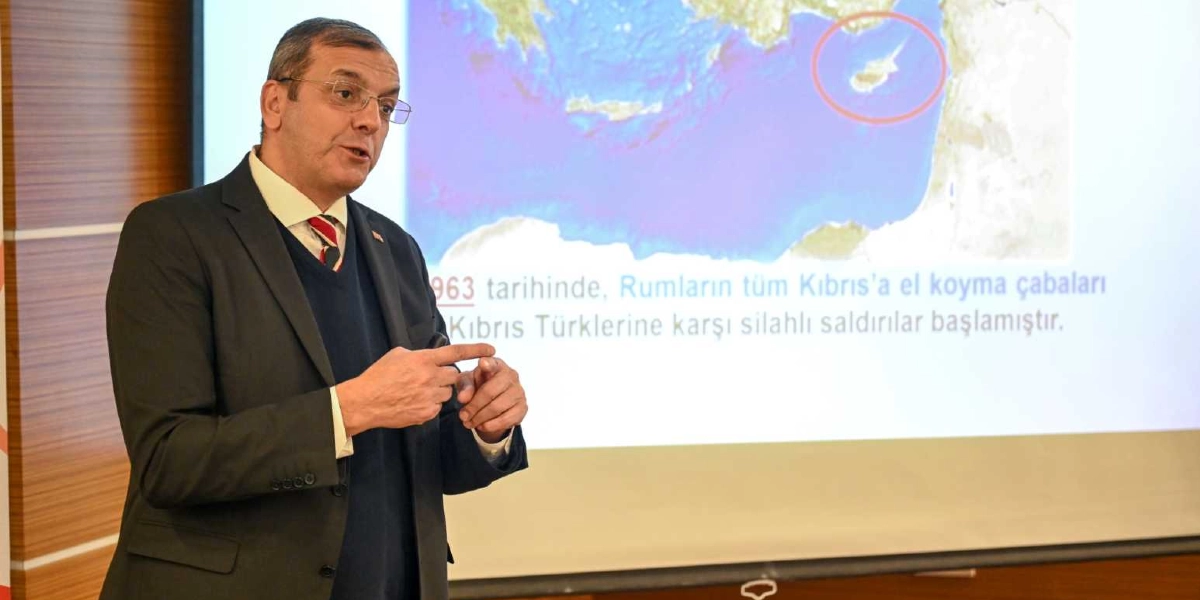 Gözlüklü, lacivert kazak ve üzerine koyu renk ceket giymiş bir erkek, arkasındaki projeksiyon perdesinde yer alan Kıbrıs haritasını işaret ederek sunum yapıyor. Perdede kırmızı bir daire içine alınmış Kıbrıs haritası ve üzerinde 