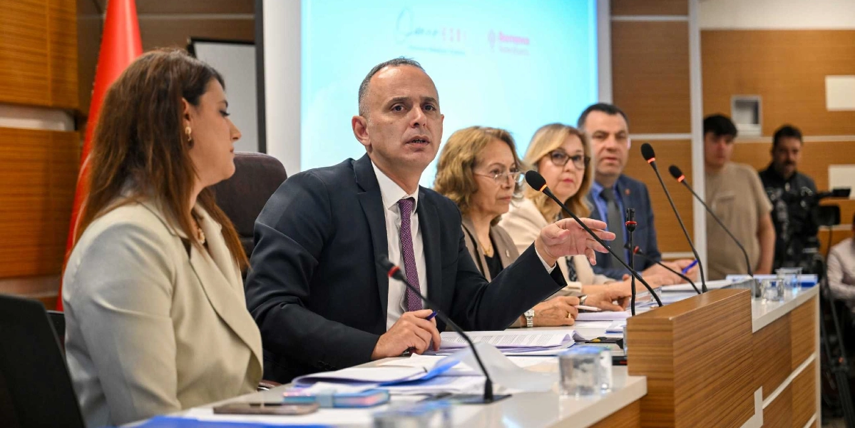 Bornova Belediyesi meclis toplantısında, lacivert takım elbiseli ve mor kravatlı bir yetkili, önündeki mikrofonla konuşma yaparken görülüyor. Masanın etrafında oturan diğer belediye meclis üyeleri kendisini dinliyor. Arka planda Bornova Belediyesi logosunun yer aldığı bir projeksiyon ekranı ve Türk bayrağı bulunuyor.