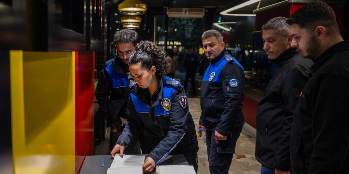 Bornova Belediyesi zabıta ekipleri, bir işletmede yılbaşı öncesi denetim gerçekleştiriyor. Üniformalı bir kadın zabıta memuru masadaki belgeleri incelerken, yanında bulunan diğer zabıta memurları ve işletme yetkilileri süreci takip ediyor.