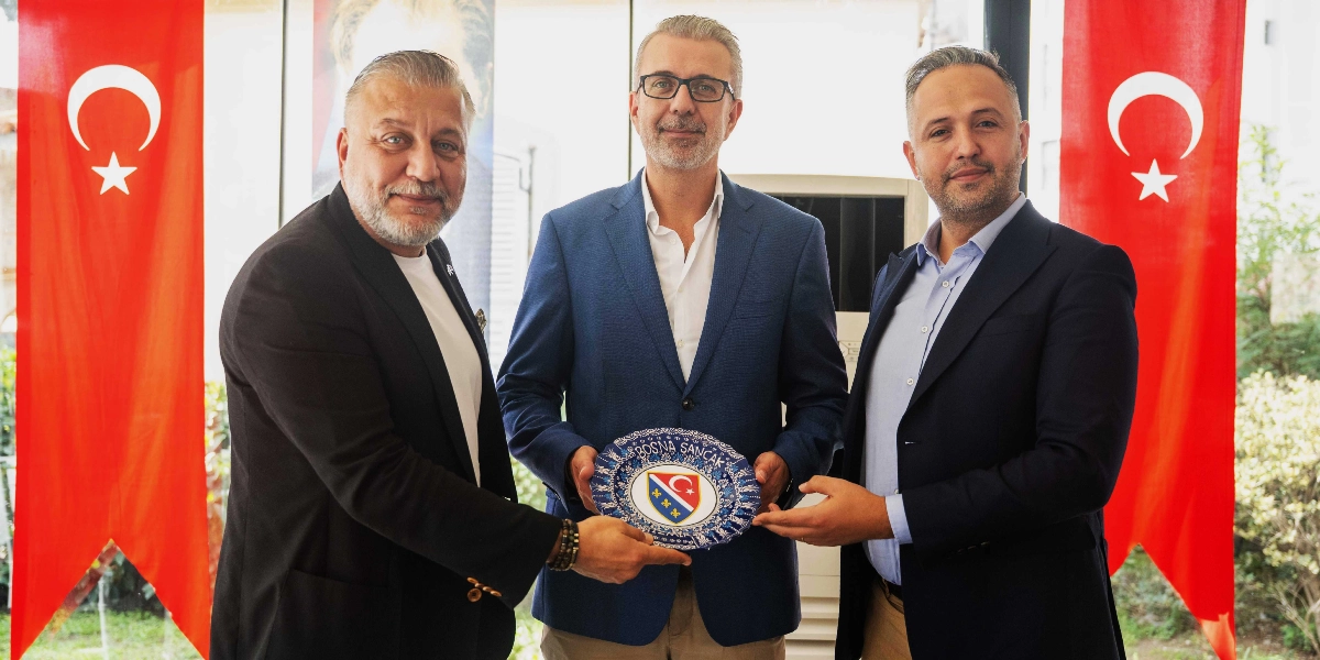 Üzerinde Bosna Sancak yazılı ve Bosna Hersek bayrağı motifli dekoratif bir tabağı birlikte tutan üç iş insanı, iki yanda asılı Türk bayraklarının önünde yan yana duruyor.