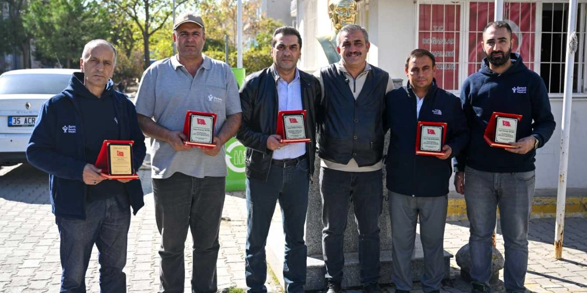 Dış mekanda, yan yana duran ve ellerinde teşekkür plaketleri tutan altı erkek. Arka planda Doğanlar Mahallesi Muhtarlığı binası, Atatürk büstü ve park halindeki bir otomobil görülüyor. Kişilerin çoğu Bornova Belediyesi logolu kıyafetler giyiyor.