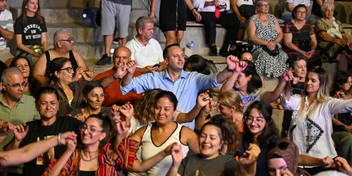 Açık havada, amfi tiyatro tarzı basamaklı oturma alanında bir araya gelmiş kalabalık bir grup insan, ellerini havaya kaldırarak coşkuyla halay çekiyor. Grubun merkezinde açık mavi gömlekli bir erkek, çevresindeki kadınlarla el ele tutuşarak neşeli bir şekilde gülümsüyor. Katılımcıların çoğu günlük kıyafetler içinde, mutlu ve enerjik bir ifadeyle kutlamaya katılıyor.