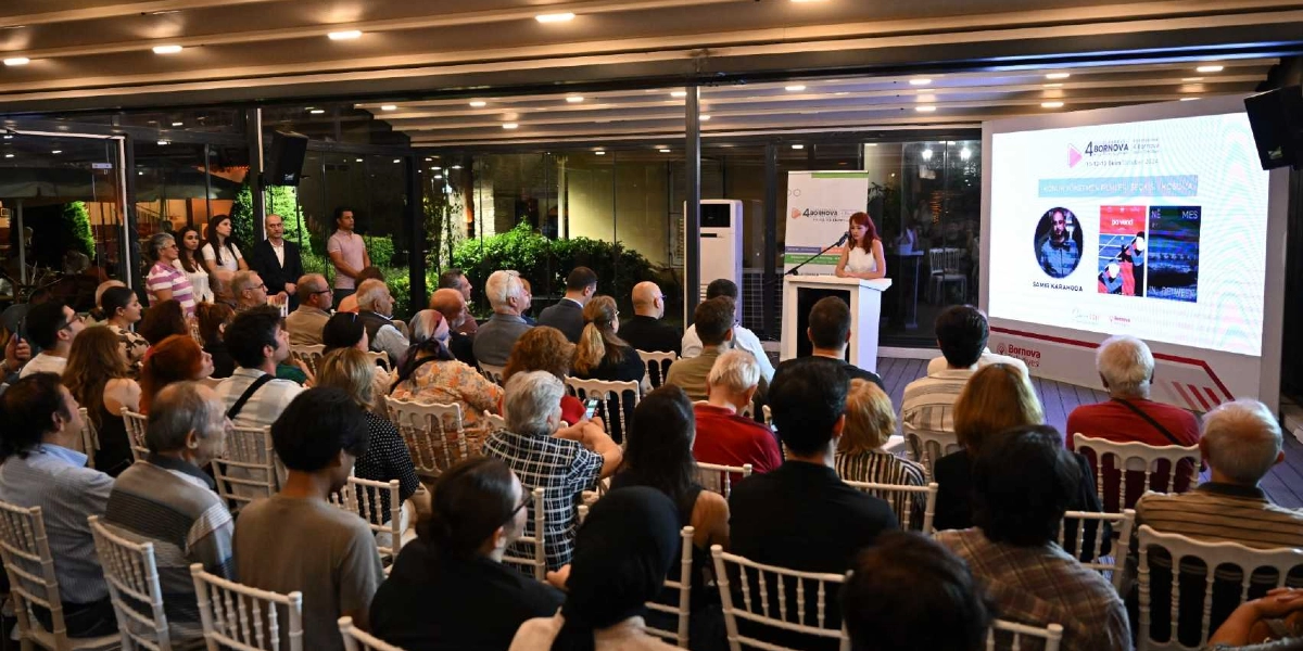 Kapalı bir mekanda düzenlenen etkinlikte, beyaz sandalyelerde oturan kalabalık bir izleyici grubu, sahnedeki konuşmacıyı ve arkasındaki dev ekranı izliyor. Ekranın üzerinde 4. Uluslararası Bornova Kısa Film Günleri logosu, konuk yönetmen Samir Karahoda'nın fotoğrafı ve film afişleri yer alıyor. Sahne kürsüsünde beyaz kıyafetli bir kadın konuşma yapıyor.
