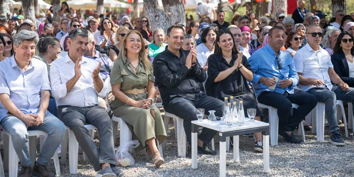 Açık havada, ağaçlık bir alanda düzenlenen etkinlikte yan yana dizilmiş beyaz sandalyelerde oturan bir grup insan. Ön sırada, aralarında Bornova Belediye Başkanı Ömer Eşki'nin de bulunduğu, alkışlayan ve gülümseyen bir grup katılımcı yer alıyor. Katılımcıların önünde üzerinde su şişeleri bulunan küçük bir masa bulunuyor. Arka planda ise etkinliği izleyen kalabalık bir topluluk ve beyaz çadırlar görülüyor.