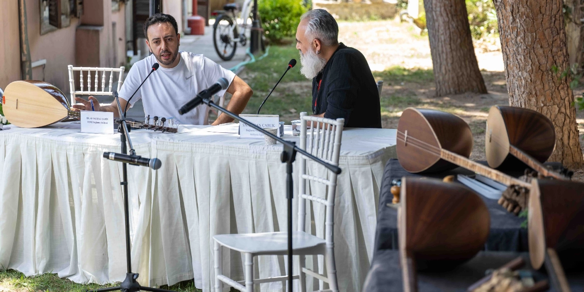 Açık havada, ağaçların altında düzenlenen bir etkinlikte, beyaz örtülü bir masanın iki yanında oturan iki erkek konuşma yapıyor. Masanın üzerinde mikrofonlar ve önlerinde isimlerinin yazılı olduğu kartlar bulunuyor. Sol tarafta oturan kişi beyaz tişörtlü, sağ tarafta oturan ise siyah kıyafetli, beyaz sakallı biridir. Masanın üzerinde ve ön taraftaki ayrı bir siyah örtülü masanın üzerinde, yapım aşamasında veya sergilenen çeşitli bağlamalar yer alıyor.