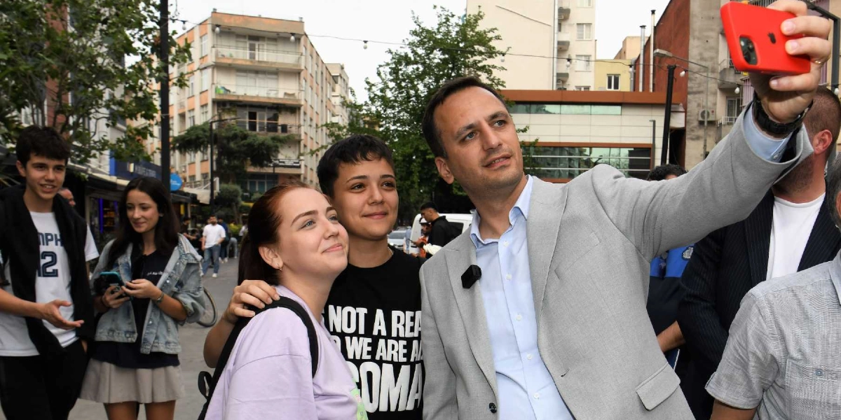 Açık renk ceketli bir erkek, Bornova sokaklarında gençlerle birlikte kırmızı kılıflı bir cep telefonuyla selfie çekiyor. Yanında duran iki genç kız gülümseyerek kameraya bakarken, arka planda farklı kıyafetler içindeki diğer gençler ve şehir dokusu yer alıyor.
