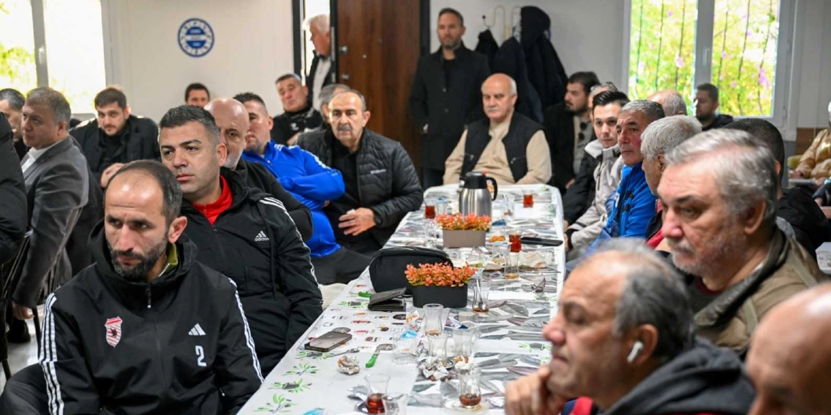 Kapalı bir mekanda, uzun bir masa etrafında toplanmış, çoğunluğu erkeklerden oluşan bir grup insan bir toplantıya katılıyor. Masanın üzerinde çay bardakları, küçük saksılarda yapay çiçekler ve bir termos bulunuyor. Katılımcıların bir kısmı spor kıyafetleri giyerken, bir kısmı günlük kıyafetlerle masada oturuyor ve dikkatle dinliyor. Arka planda duvarda mavi beyaz renkli bir spor kulübü amblemi yer alıyor.