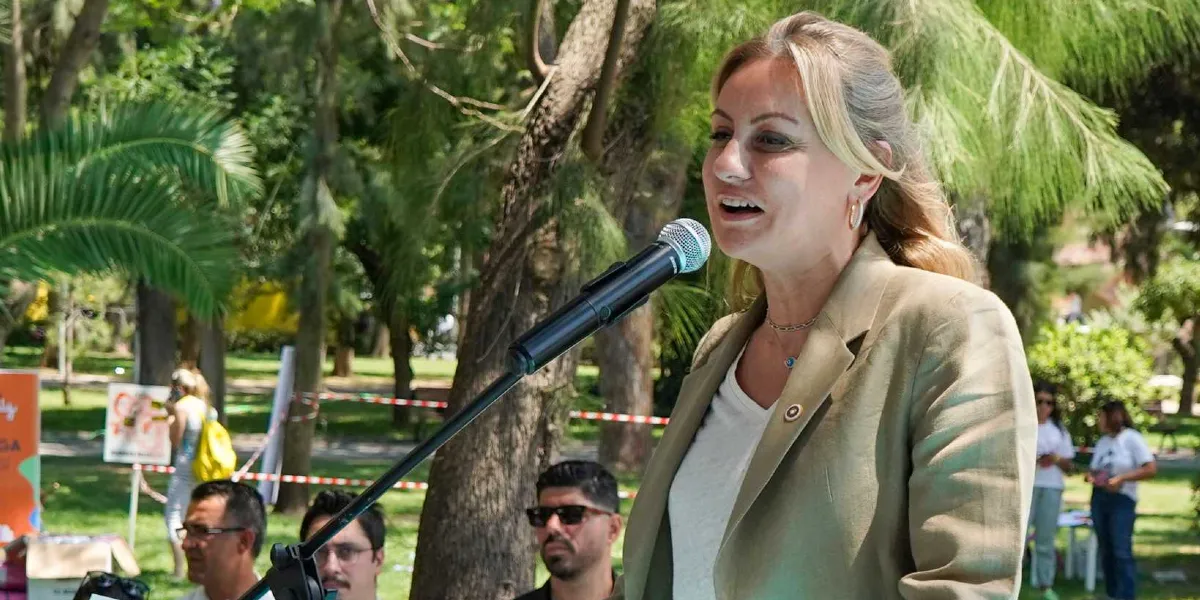 Açık havada, ağaçlık bir alanda düzenlenen etkinlikte konuşma yapan sarı saçlı bir kadın. Kadın, bej rengi bir ceket ve beyaz bir tişört giymiş, mikrofon başında konuşuyor. Arka planda ise etkinliğe katılan diğer insanlar ve ağaçlar görülüyor.