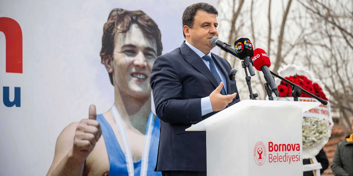 Lacivert takım elbiseli ve mavi kravatlı bir yetkili, üzerinde Bornova Belediyesi logosu bulunan bir kürsüde mikrofon başında konuşma yapıyor. Arka planda, halterci Naim Süleymanoğlu'nun gençlik yıllarına ait, baş parmağıyla 