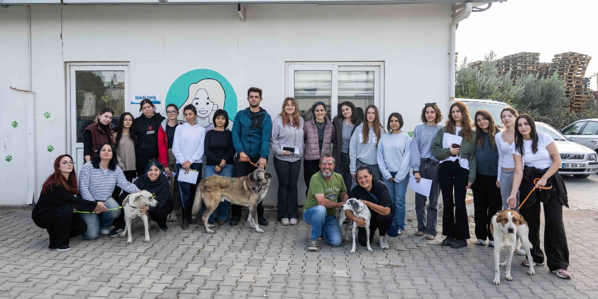 Beyaz bir binanın önünde yan yana duran, aralarında köpeklerin de bulunduğu kalabalık bir grup genç ve yetişkin. Binanın duvarında 