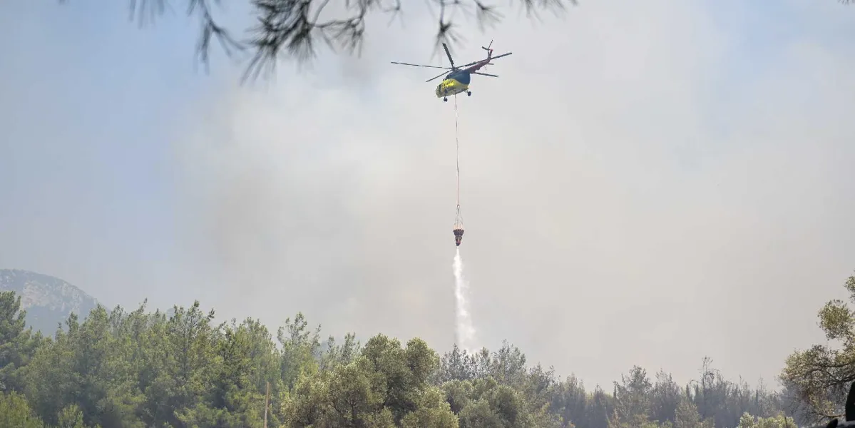Sarı ve kırmızı renkli bir yangın söndürme helikopteri, ormanlık alanın üzerinde havada asılı kalarak altındaki bambi kovasından aşağıya su boşaltıyor. Arka planda dumanla kaplı bir gökyüzü ve ön planda yeşil ağaçlar görülüyor.