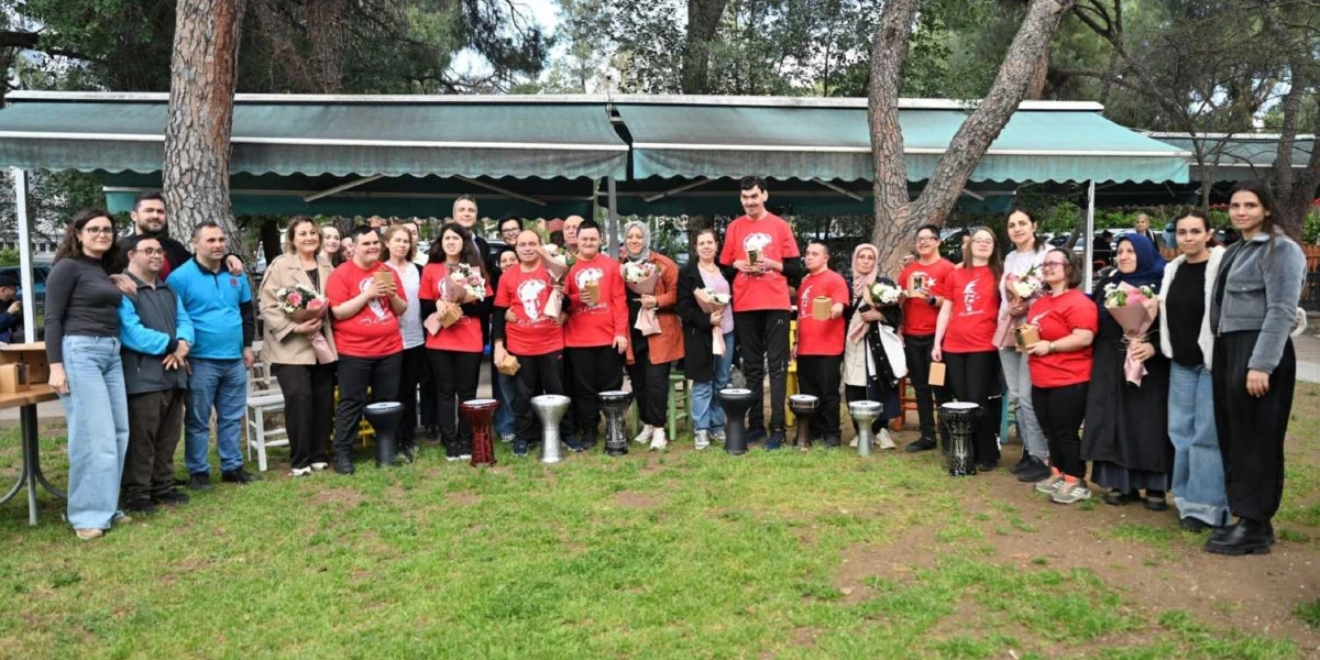 Açık havada, ağaçların altında ve yeşil bir alanda bir araya gelmiş, aralarında Down sendromlu bireylerin de bulunduğu kalabalık bir grup insan yan yana durarak poz veriyor. Grubun büyük bir kısmı üzerinde Atatürk silüeti bulunan kırmızı tişörtler giyiyor ve ellerinde çiçek buketleri ile küçük hediye kutuları tutuyor. Bazı kişilerin önünde yere dizilmiş darbuka benzeri müzik aletleri bulunuyor. Arka planda yeşil bir tente ile kaplı bir kafe alanı ve ağaçlar yer alıyor.