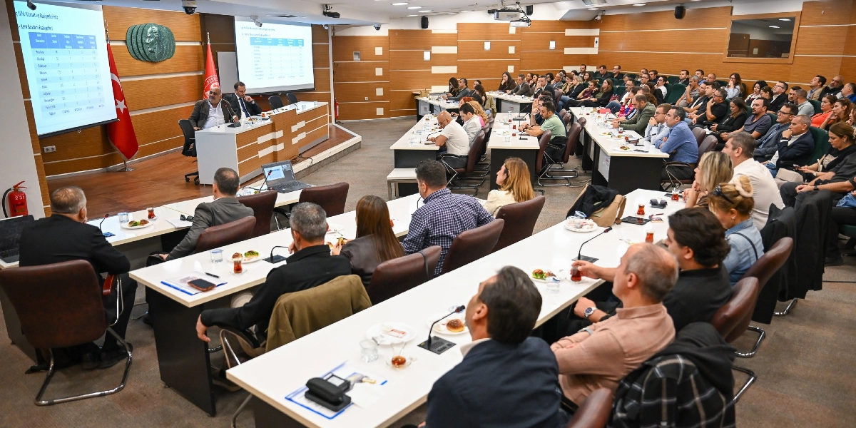 Modern bir toplantı salonunda, projeksiyon ekranında Bornova Belediyesi'nin hizmet ve faaliyet verilerinin yer aldığı bir sunumun yapıldığı toplantıya katılan çok sayıda kişi. Salonun ön kısmında konuşmacıların bulunduğu bir kürsü, arkasında Türk bayrağı ve Atatürk rölyefi yer alırken, katılımcılar uzun masalarda oturarak sunumu takip ediyor.
