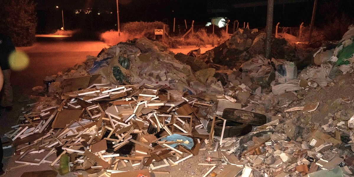 Gece vakti boş bir arazide, yol kenarına yığılmış büyük miktarda inşaat atığı, moloz, ahşap çerçeve parçaları ve eski araba lastikleri görülüyor.