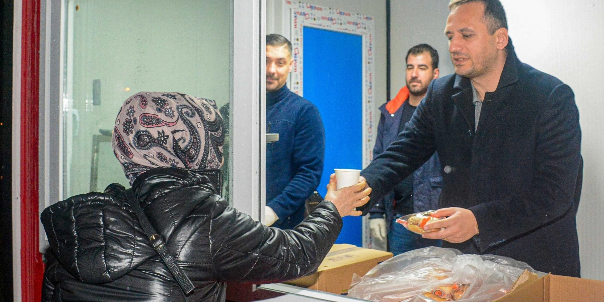 Siyah şişme montlu ve desenli başörtülü bir kadın, bir pencerenin önünde durarak içerideki görevliden bir bardak sıcak içecek ve paketli bir yiyecek alıyor. Pencerenin arkasında, koyu renkli kaban giymiş bir erkek, kadına ikramda bulunurken, yanında iki erkek daha bekliyor. Önlerinde içinde paketli yiyeceklerin bulunduğu karton kutular yer alıyor.