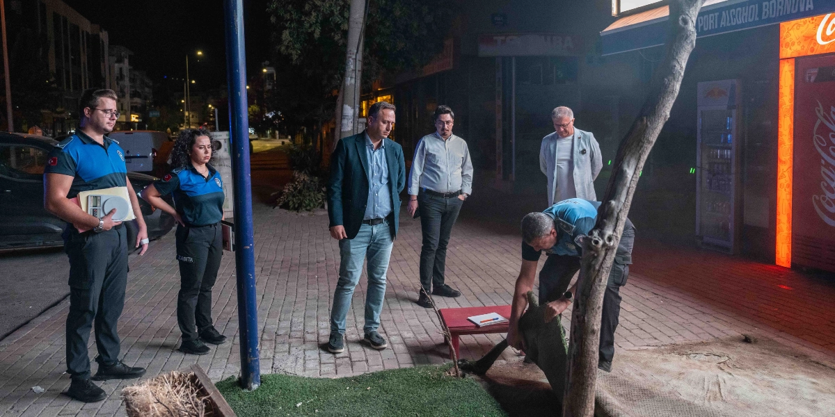 Gece vakti bir kaldırım üzerinde zabıta ekipleri ve sivil kıyafetli yetkililer denetim yapıyor. Üniformalı bir zabıta memuru eğilerek yerdeki bir örtüyü kaldırırken, diğer iki zabıta memuru ve üç sivil yetkili onları izliyor. Arka planda aydınlatılmış bir dükkan girişi ve Coca-Cola tabelası görülüyor.