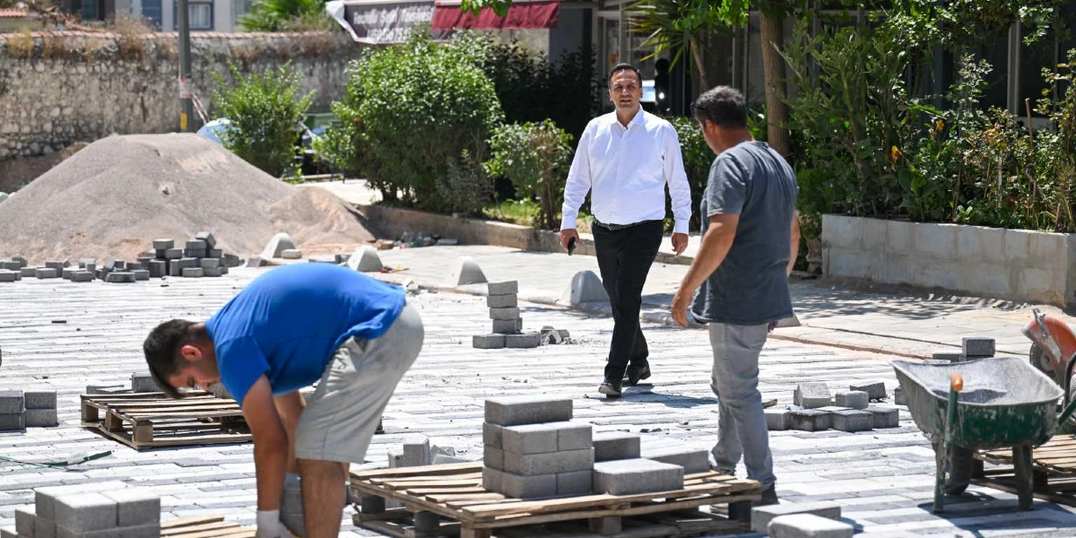 Bornova'da devam eden bir çevre düzenleme çalışmasında, mavi tişörtlü bir işçi yerdeki parke taşlarını döşerken, yanında gri tişörtlü bir başka işçi ve beyaz gömlekli bir yetkili çalışmaları incelemektedir. Çalışma alanında yığın halinde kum, ahşap paletler üzerinde parke taşları ve bir el arabası bulunmaktadır.