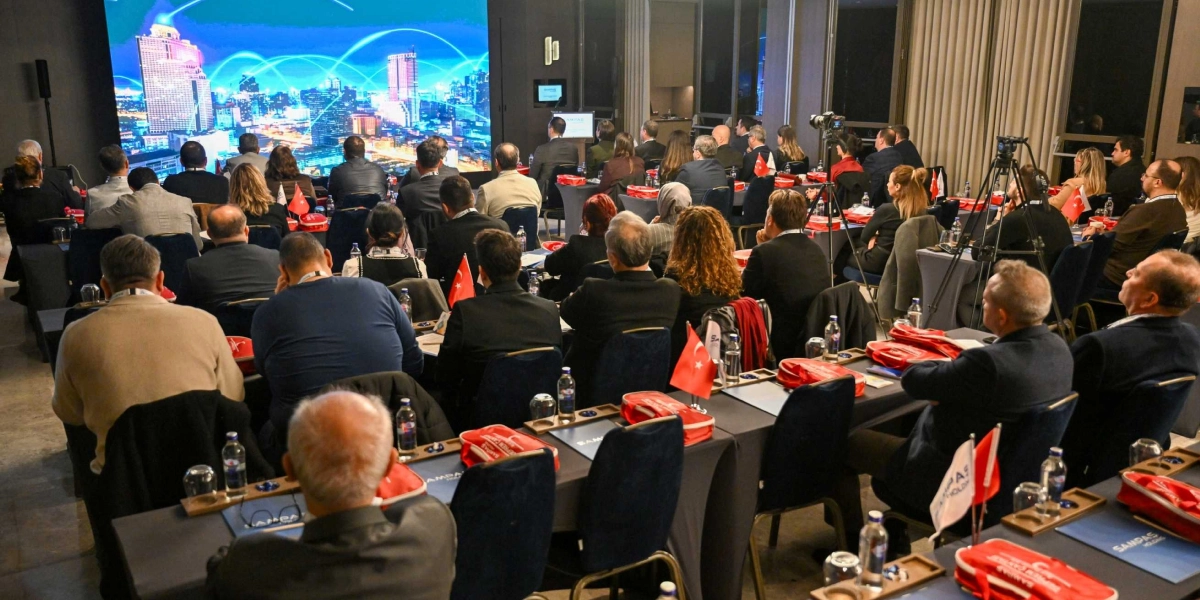 Modern bir konferans salonunda, dijital ağ bağlantılarını temsil eden grafiklerin yer aldığı dev bir ekrana dönük oturan kalabalık bir dinleyici grubu. Katılımcılar masalarda oturuyor, önlerinde Türk bayrakları, su şişeleri ve üzerinde kurum logoları bulunan kırmızı çantalar yer alıyor. Salonun arka kısmında profesyonel kamera ekipmanları bulunuyor ve ortamda resmi bir toplantı havası hakim.