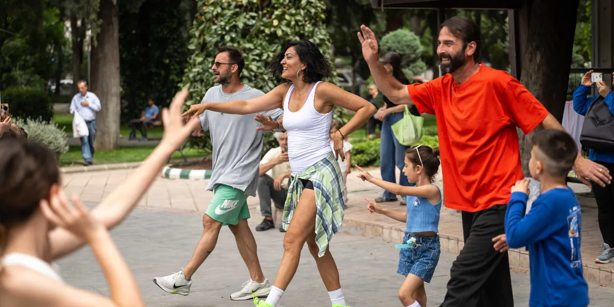 Açık havada, park benzeri bir alanda neşeyle dans eden bir grup yetişkin ve çocuk. Ön planda turuncu tişörtlü bir adam, beyaz atletli bir kadın ve gri tişörtlü bir adam müzik eşliğinde hareket ediyor. Kadının beline bağladığı ekose bir gömlek bulunuyor. Yanlarında mavi kıyafetli bir kız çocuğu ve bir erkek çocuk da onlara eşlik ediyor. Arka planda ise ağaçlar, bankta oturan insanlar ve yürüyen kişiler yer alıyor.