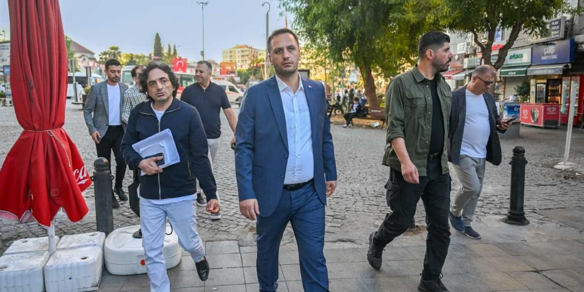 Bornova'da bir meydanda, aralarında Bornova Belediye Başkanı Ömer Eşki'nin de bulunduğu bir grup erkek, kaldırımda yürüyerek ilerliyor. Ömer Eşki, lacivert takım elbisesi ve beyaz gömleğiyle grubun önünde yer alıyor. Yanında koyu renkli bir ceket ve beyaz pantolon giyen, elinde evraklar tutan bir kişi ile haki renkli bir ceket ve pantolon giyen başka bir kişi bulunuyor. Grubun arkasında ise farklı kıyafetlerle yürüyen diğer kişiler yer alıyor. Arka planda ağaçlar, dükkan tabelaları ve bir dondurma dolabı görünen meydan, sabah saatlerinin aydınlığında hareketli bir görünüm sergiliyor.