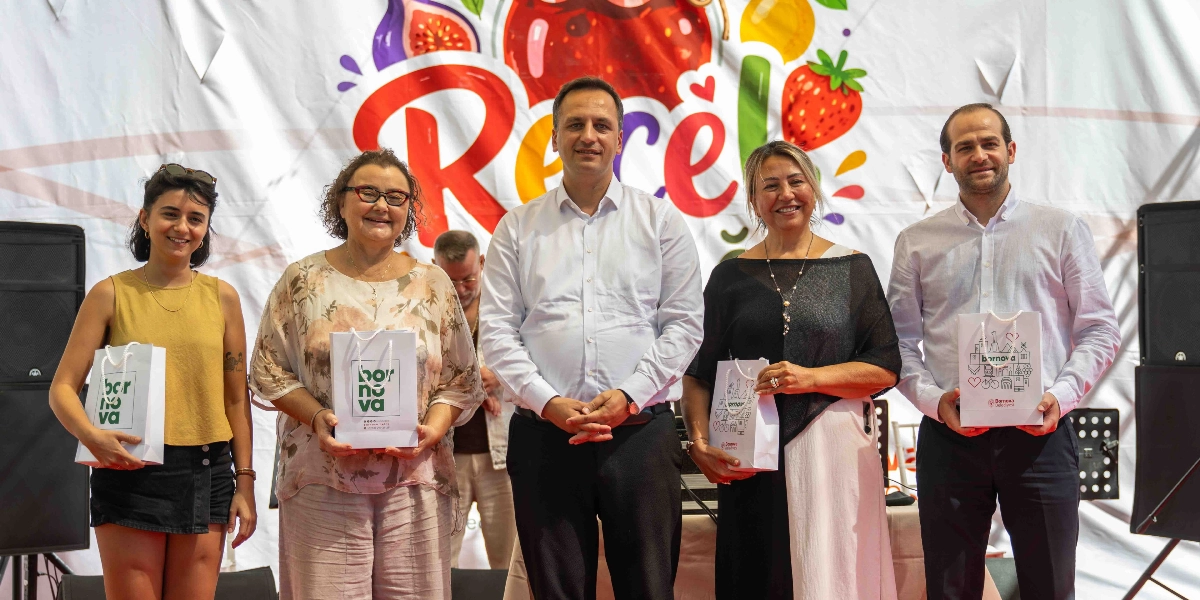 Üzerinde renkli meyve çizimleri ve büyük harflerle Reçel yazan bir pankartın önünde duran beş kişi, ellerinde Bornova Belediyesi logolu hediye çantalarıyla objektiflere gülümsüyor. Soldan sağa doğru; sarı kolsuz bluz ve siyah şort giyen genç bir kadın, çiçek desenli bluz ve açık renkli pantolon giyen orta yaşlı bir kadın, beyaz gömlek ve siyah pantolon giyen bir erkek, siyah bluz ve uzun etek giyen bir kadın ile yine beyaz gömlek ve siyah pantolon giyen bir erkek yer alıyor.