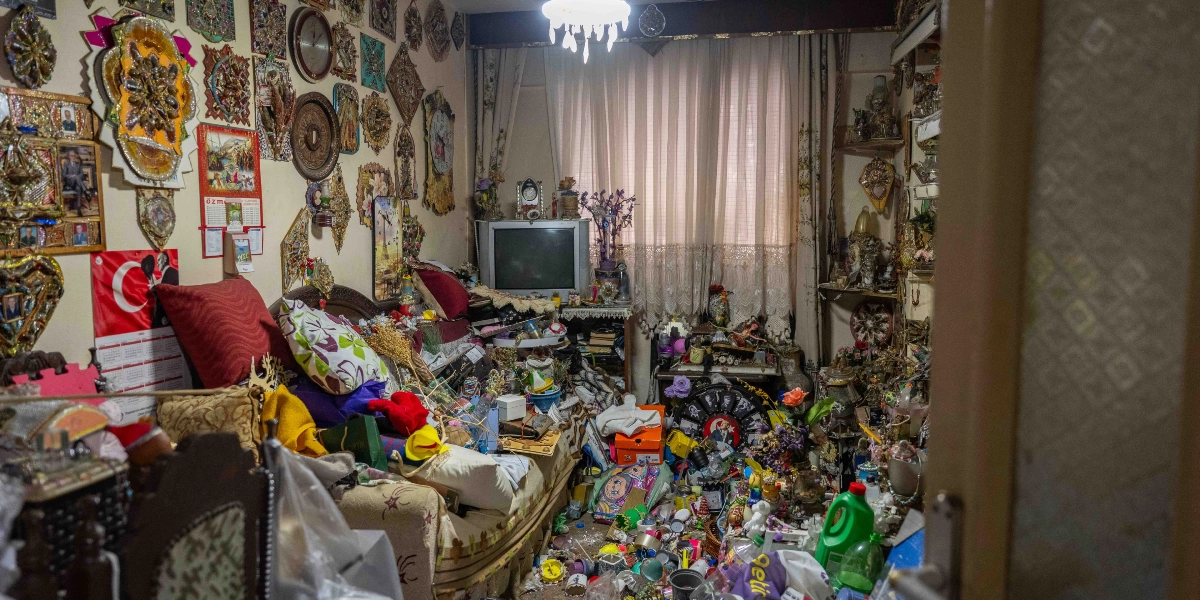 Duvarları çok sayıda süs eşyası, saat ve tabloyla kaplı, eşyalarla aşırı derecede dolu ve dağınık bir oturma odası. Odanın ortasındaki koltukların üzeri yastıklar, kutular ve çeşitli objelerle yığılmış durumdayken, zemin ise plastik şişeler, temizlik malzemeleri ve çok sayıda küçük eşyadan oluşan bir yığınla kaplı. Pencere önünde eski tip bir televizyon ve raflarda yoğun bir şekilde biriktirilmiş dekoratif objeler göze çarpıyor.