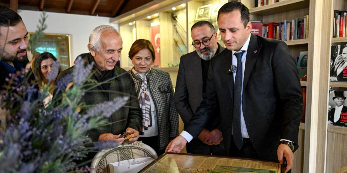 İç mekanda, kitaplıklarla çevrili bir alanda bir araya gelen beş kişilik bir grup, masanın üzerindeki tarihi bir haritayı inceliyor. Takım elbiseli ve kravatlı bir erkek, haritayı işaret ederek yanındaki kişilere bilgi veriyor. Grubun solunda, elinde küçük bir nesne tutan beyaz saçlı bir beyefendi, yanında bir kadın ve diğer katılımcılar dikkatle haritaya bakıyor. Arka planda raflarda kitaplar ve Atatürk fotoğrafları yer alıyor.