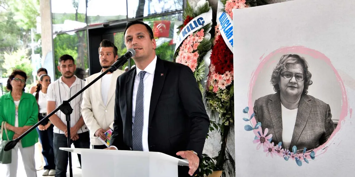 Bornova Belediye Başkanı Mustafa İduğ, kürsüde konuşma yaparken; yanında onurlandırılan emektar bürokratın siyah beyaz bir portresinin yer aldığı pano ve çiçek aranjmanları bulunuyor. Arka planda ise törene katılan diğer kişiler yer alıyor.