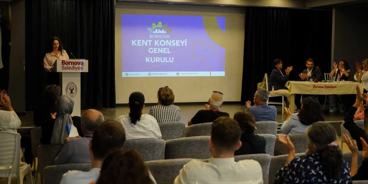 Bornova Belediyesi'nin düzenlediği bir toplantıda, kürsüde konuşma yapan bir kadın ve onu dinleyen katılımcılar yer alıyor. Salonun ön tarafındaki projeksiyon perdesinde 