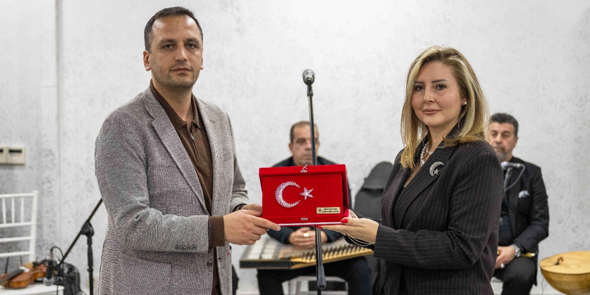 Gri ceketli bir erkek ve siyah çizgili ceketli bir kadın, üzerinde ay yıldız işlemesi bulunan kırmızı kadife bir plaketi birlikte tutuyor. Arka planda, kanun çalan bir müzisyen ve yanında oturan başka bir kişi daha bulunuyor.