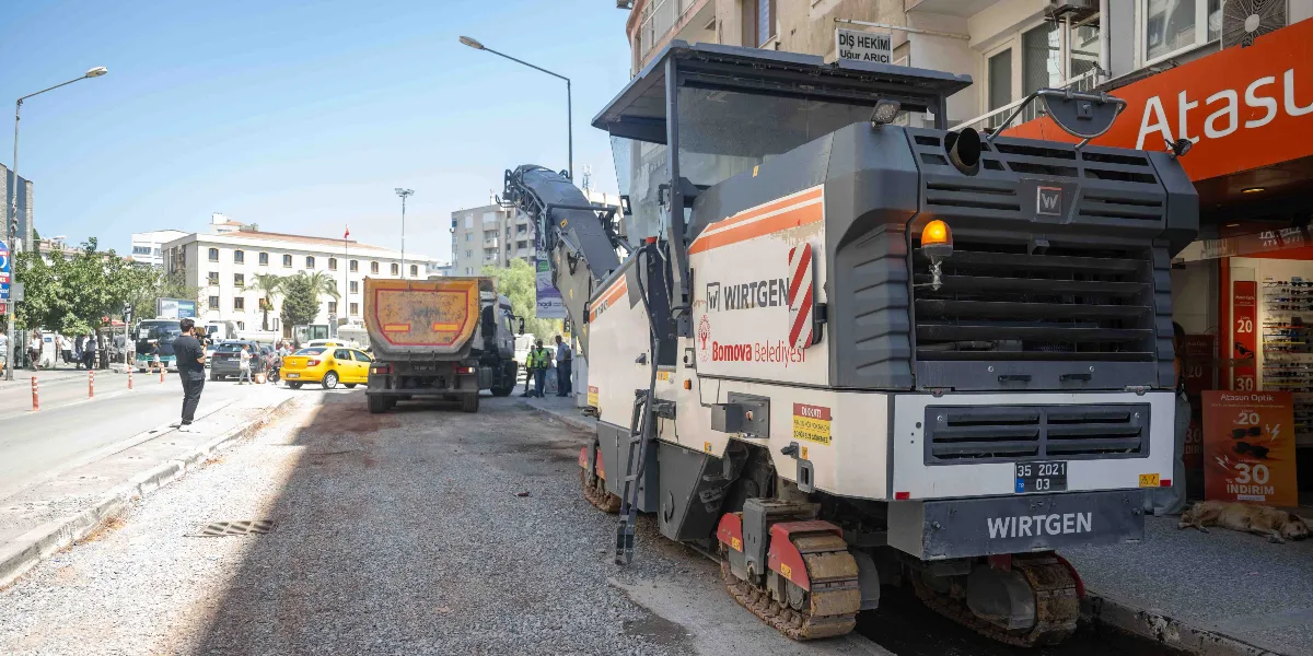 Bornova Belediyesi'ne ait Wirtgen marka paletli asfalt kazıma makinesi, Mustafa Kemal Caddesi'ndeki yol yenileme çalışmaları kapsamında asfaltı kazırken görülüyor. Makinenin önünde bir hafriyat kamyonu beklerken, kaldırımda Atasun Optik mağazası ve çevredeki yayalar yer alıyor.