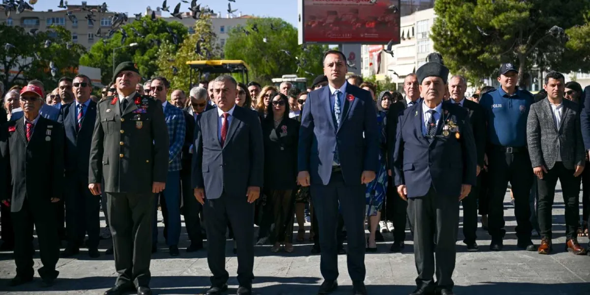 Gaziler Günü töreninde, aralarında askeri üniformalı bir subay, madalyalı gaziler, Bornova Belediye Başkanı ve diğer protokol üyelerinin bulunduğu bir grup insan, açık havada yan yana durarak saygı duruşunda bulunuyor. Arka planda uçuşan güvercinler ve üzerinde 